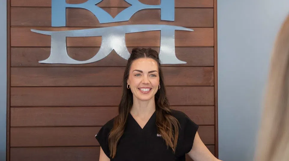 Smiling woman in black medical scrubs stands against a wooden wall with a large stylized logo behind her.