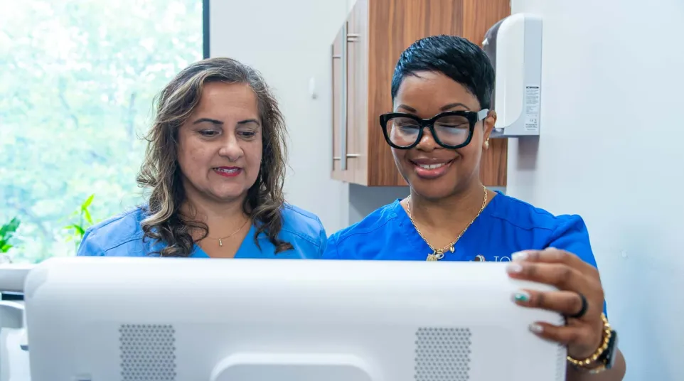 Two female healthcare professionals in blue scrubs looking at a computer screen in a medical office.