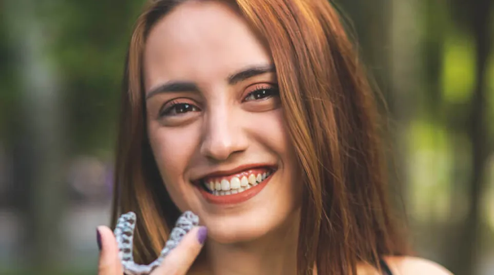 Smiling woman with red hair holding a clear orthodontic aligner outdoors with blurred green background