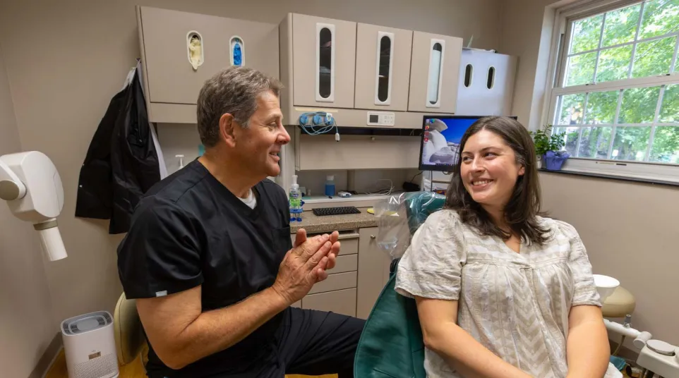 Dentist in black scrubs talking with female patient smiling in dental office with equipment and window light.