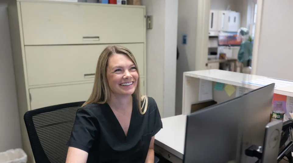 Smiling female medical receptionist in black scrubs sitting at a desk with computer in a medical office.