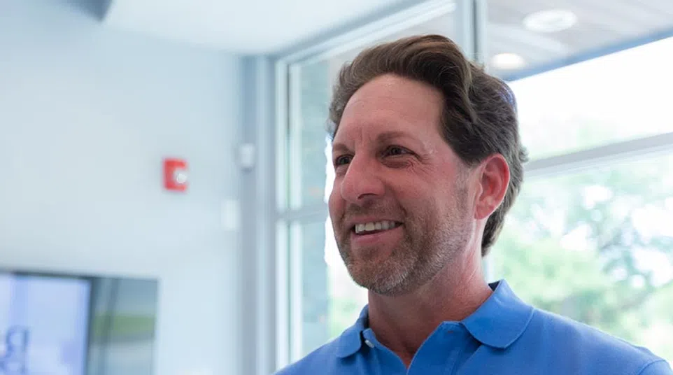 Smiling man in a blue polo shirt standing indoors near large windows with natural light.
