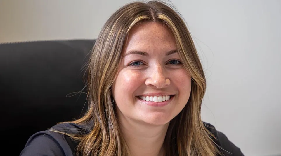 Smiling young woman with shoulder-length hair sitting at a desk in front of a laptop in an office setting.