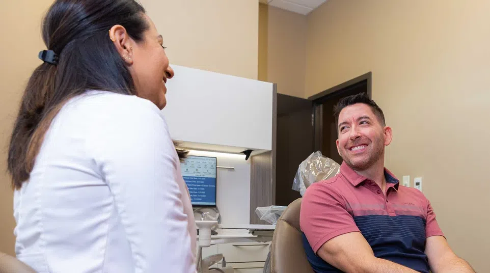 Male patient sitting in dental chair smiling and talking with female dentist in clinic room.