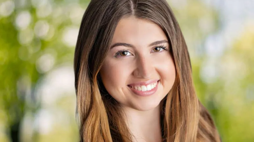 Young woman with long brown hair smiling outdoors with blurred green background wearing a black shirt
