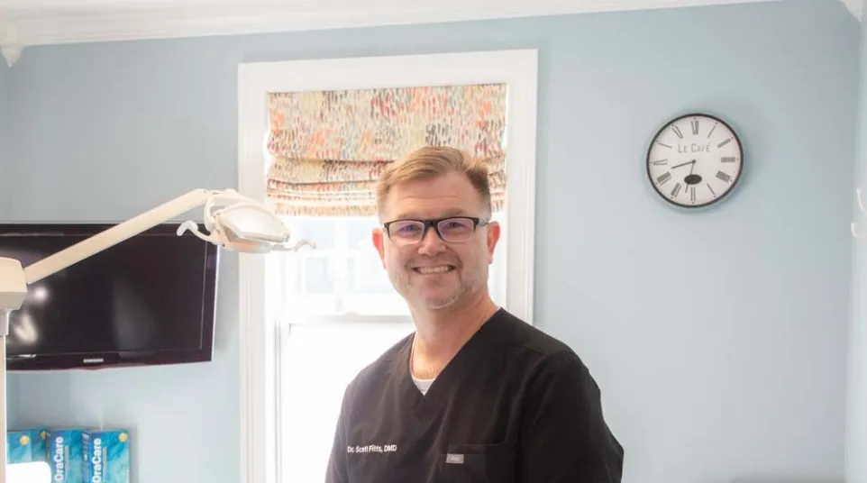 Dentist in black scrubs smiling in a bright dental office with equipment and a wall clock.