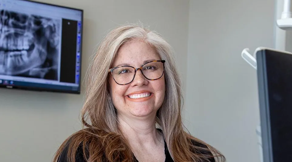 Smiling female dentist with glasses in black scrubs sitting in dental office with dental X-ray on screen behind her