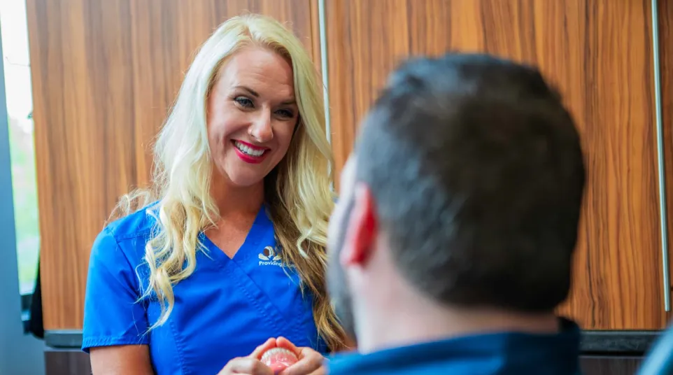 Female dental professional in blue scrubs explaining dentures to a male patient in a clinic.