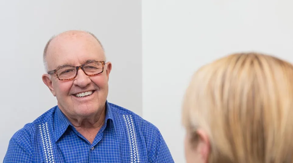 Smiling elderly man in blue checkered shirt talking to a blonde woman in a casual indoor setting.