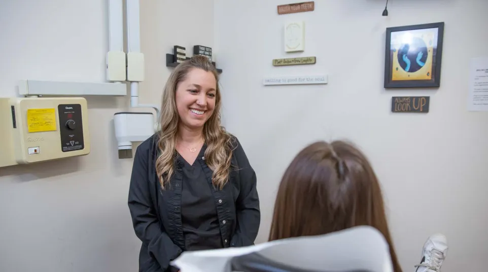 Dental hygienist smiling and talking to patient seated in dental chair in a clean office room.