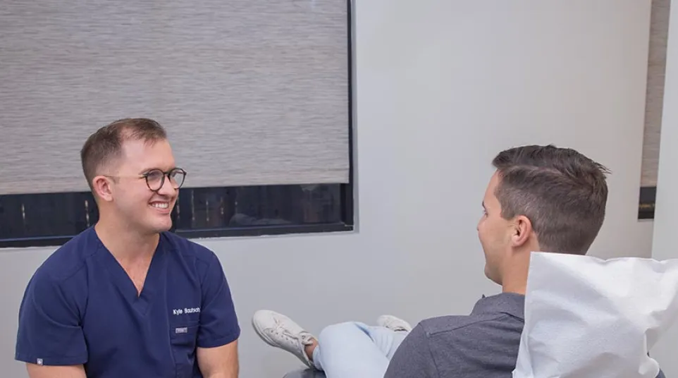 A dentist in navy scrubs smiling and talking to a male patient seated in a dental chair during consultation.