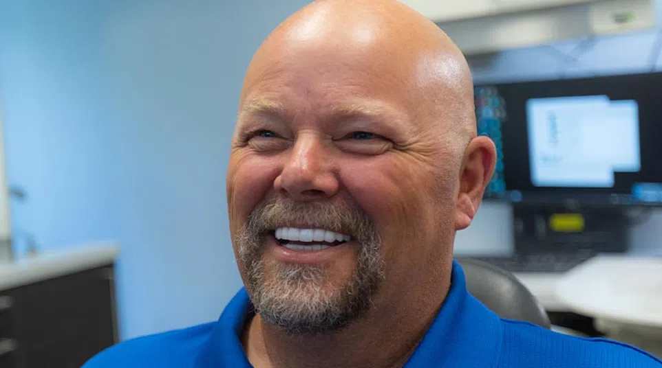 Smiling bald man with beard wearing a blue polo shirt sitting in an office with computer screens in the background