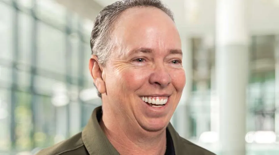 Middle-aged man in olive green Under Armour polo shirt smiling in bright modern indoor space.