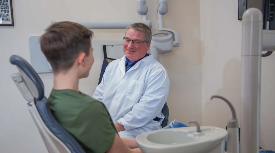 Dentist in white coat smiling while consulting a young male patient in a dental office.