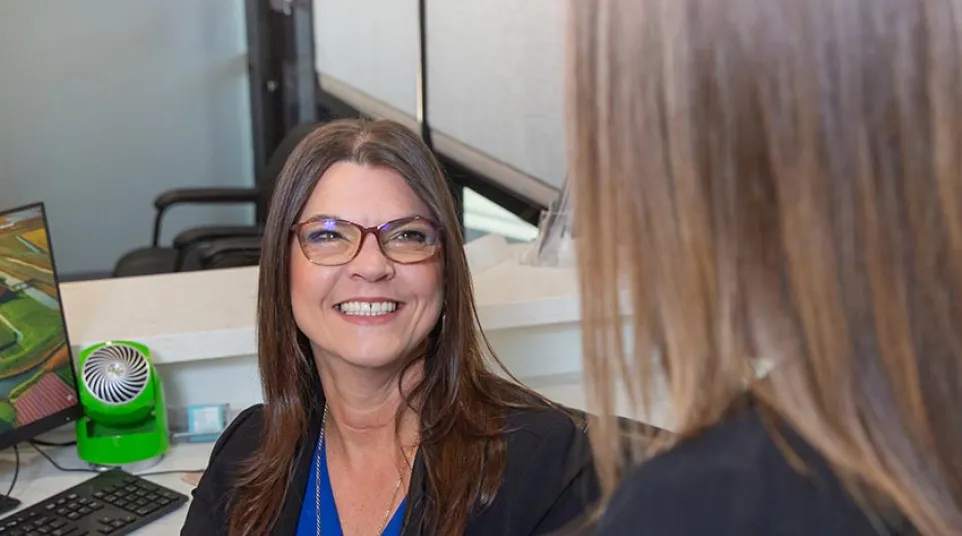 Smiling woman with glasses and blue blouse consulting another person at office desk with computer.