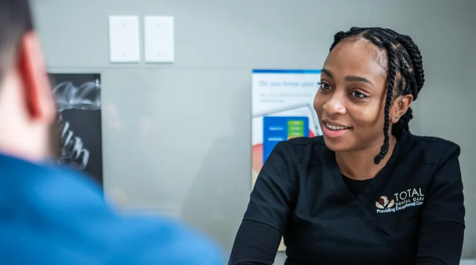 Smiling dental professional in black Total Dental Care uniform consulting a patient in a clinic setting.