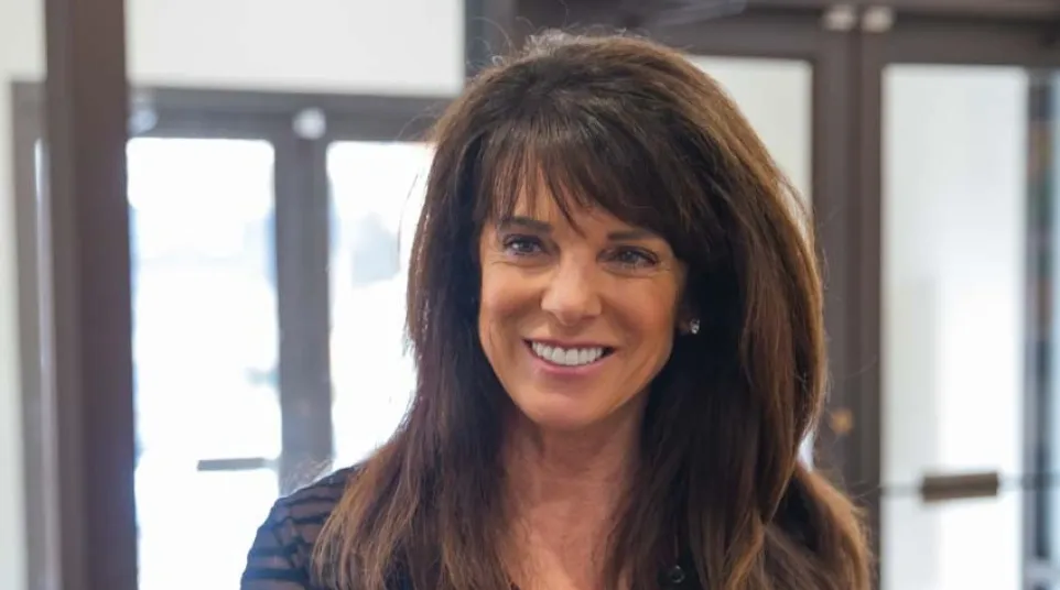 Smiling woman with brown hair wearing a maroon top and black cardigan standing indoors near a door.