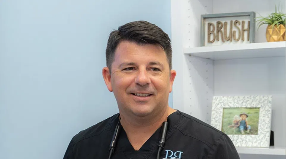 Smiling male dentist in black scrubs with magnifying glasses around neck in dental office setting.