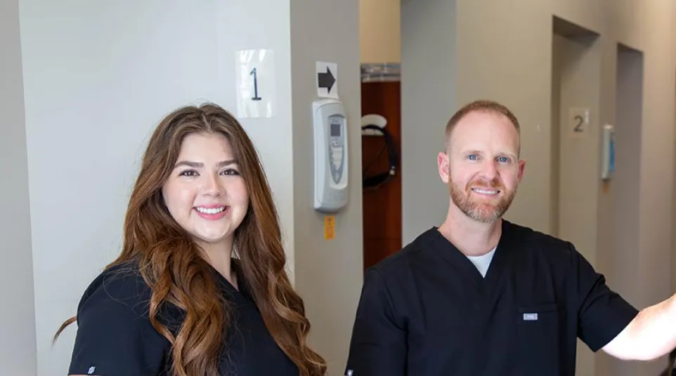 Two healthcare professionals in black scrubs smiling in a medical clinic hallway with numbered rooms.