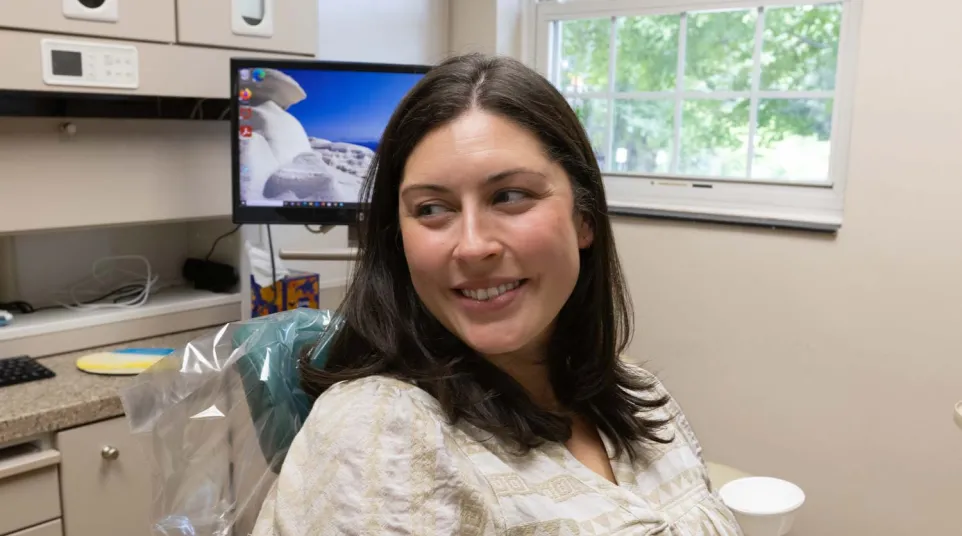 Smiling woman sitting in a dental chair in a bright dental office with computer and window in background
