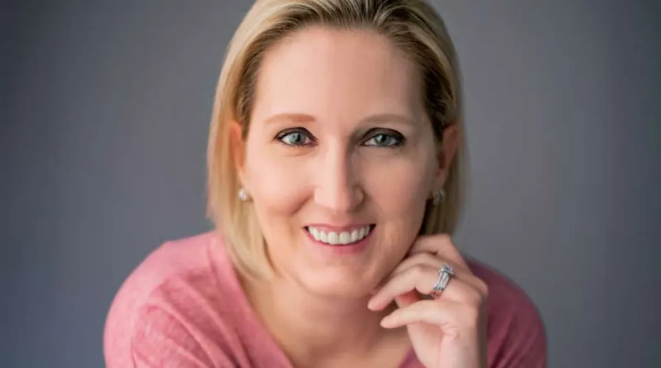 Smiling woman with short blonde hair wearing a pink top and smartwatch against a gray background.