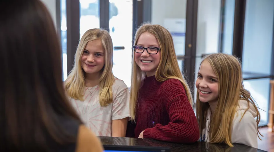 Three smiling teenage girls at a reception desk interacting with a staff member indoors in a bright setting.
