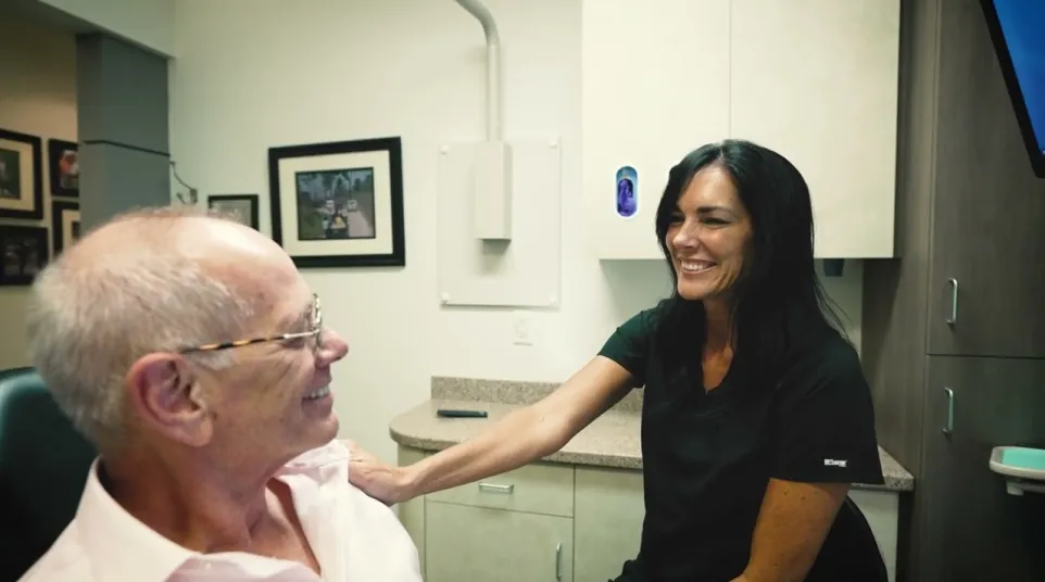 Smiling healthcare worker comforting elderly man in a medical office with framed pictures on the wall.