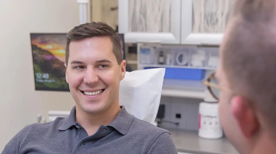 Smiling young man in a gray polo shirt sitting in a dental office chair talking to a dentist.