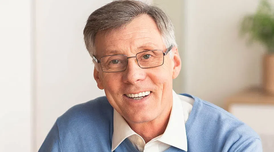 Smiling mature man wearing glasses and a blue sweater sitting indoors with a blurred background