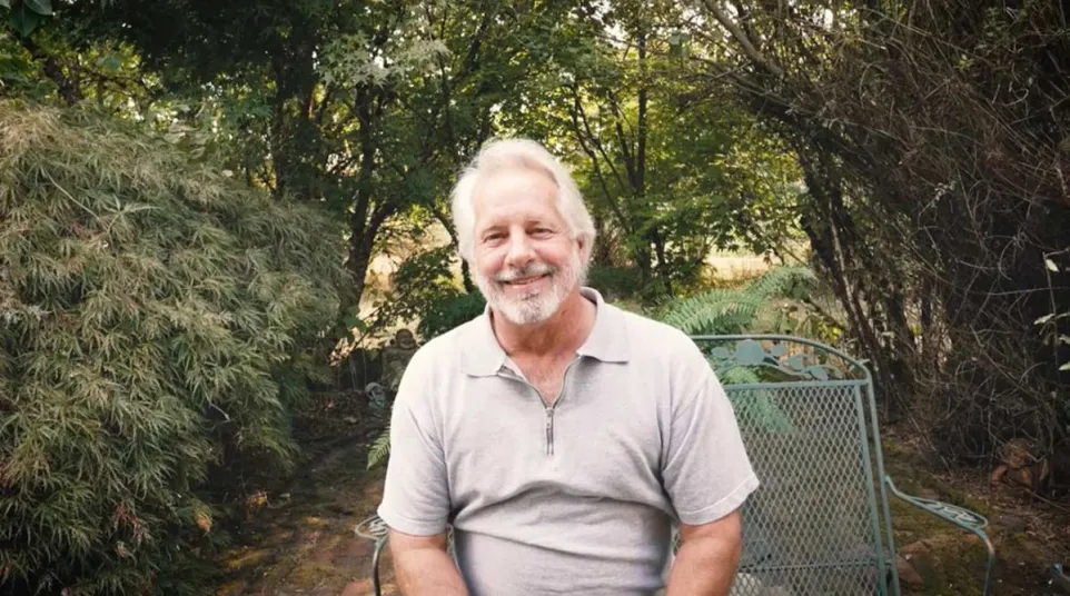 Smiling elderly man with white hair sitting on a metal chair in a lush green garden outdoors.