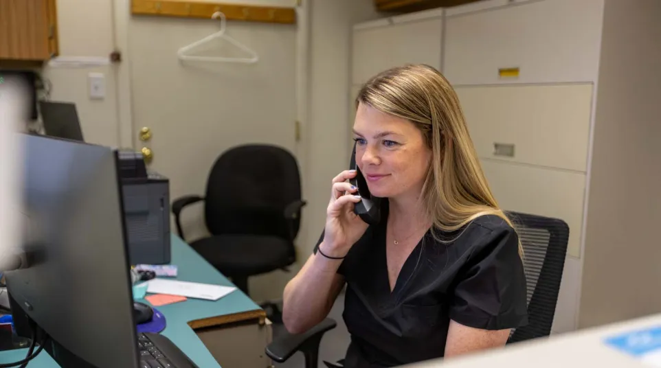 Woman in black scrubs sitting at desk using phone and computer in an office setting.