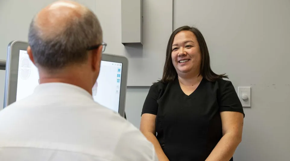 Smiling female healthcare professional talks with senior male patient in medical office with computer screen.