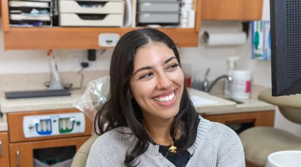 Smiling woman sitting in a dental office chair with cabinets and medical equipment in the background.