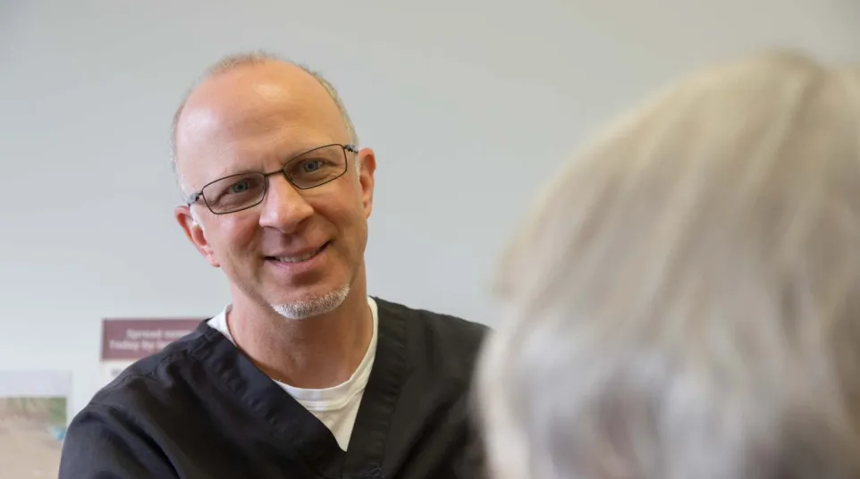 Smiling male healthcare professional in black scrubs talking to a patient with gray hair in a clinical setting.