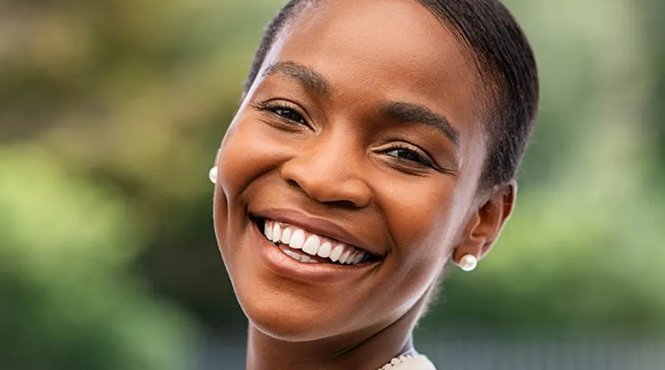 Smiling young woman with smooth skin and pearl earrings in a bright outdoor setting.