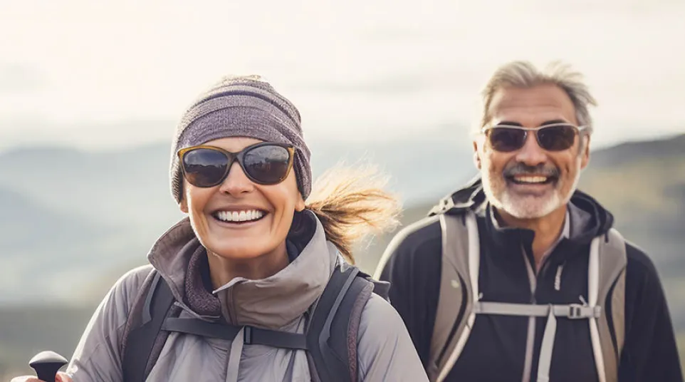 Smiling middle-aged couple hiking outdoors wearing sunglasses and backpacks with trekking poles in a mountainous area.
