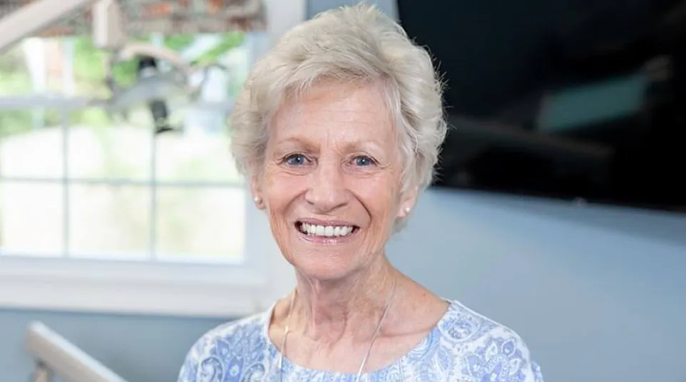 Smiling elderly woman with short white hair wearing a blue patterned top and cross necklace indoors