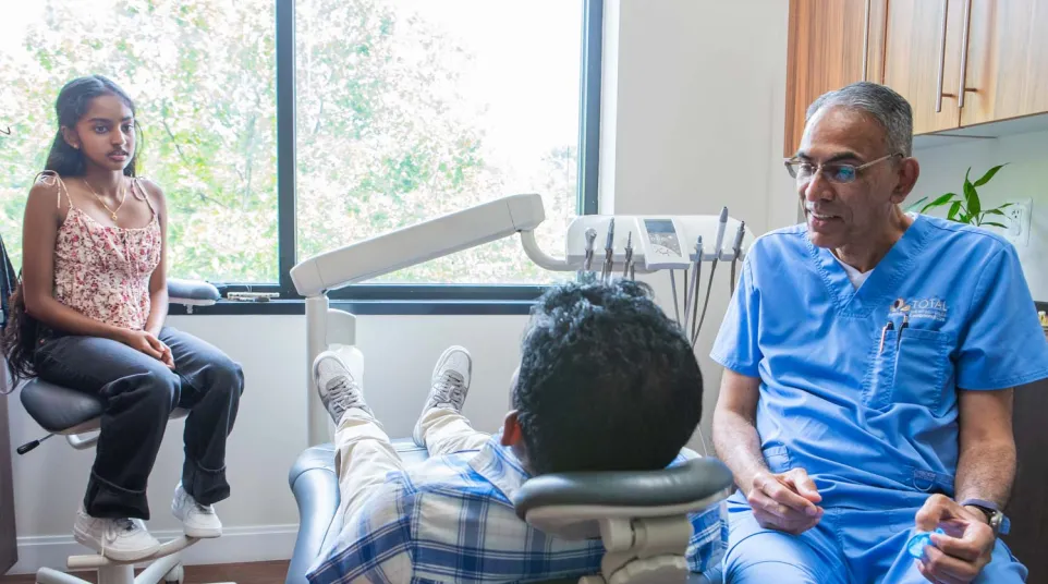 Dentist in blue scrubs talking to a boy in a dental chair with a girl waiting nearby in a bright clinic.