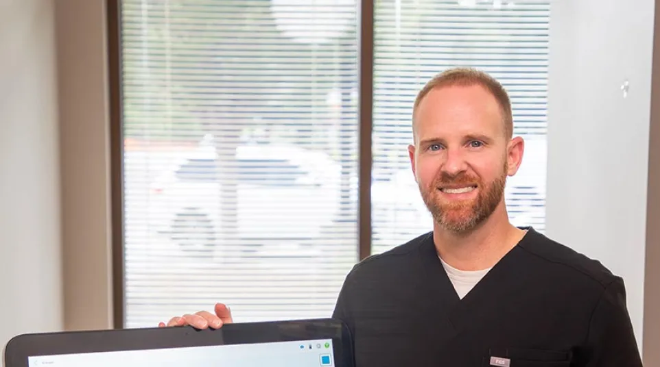 Male dentist in black scrubs displaying digital 3D dental scan on screen in bright office with window blinds.