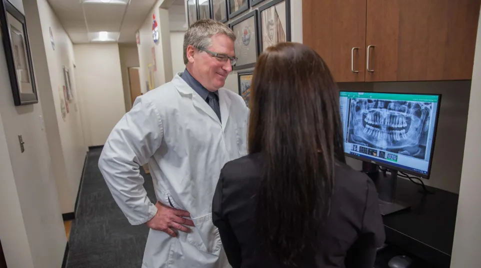 Dentist and patient reviewing a dental X-ray image on a computer screen in a clinic hallway.