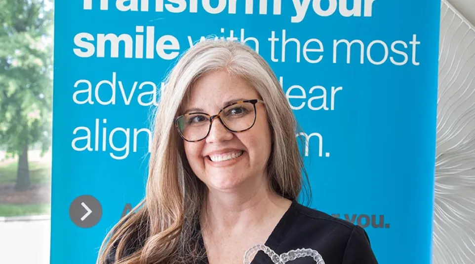 Smiling woman in glasses holding clear dental aligners shaped like a heart in front of a blue orthodontic sign.