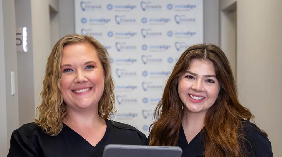 Two smiling female dental professionals in black scrubs holding a tablet in a dental clinic hallway.
