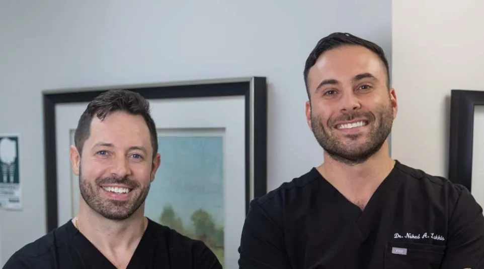 Two male dentists in black scrubs smiling with arms crossed in a dental office setting