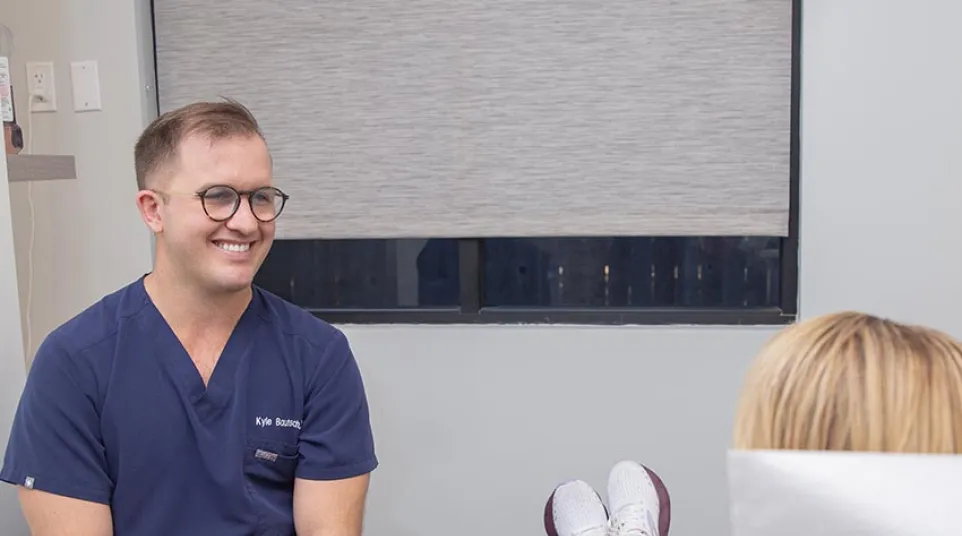 Male healthcare professional in navy scrubs smiling and talking to patient reclining on medical chair.