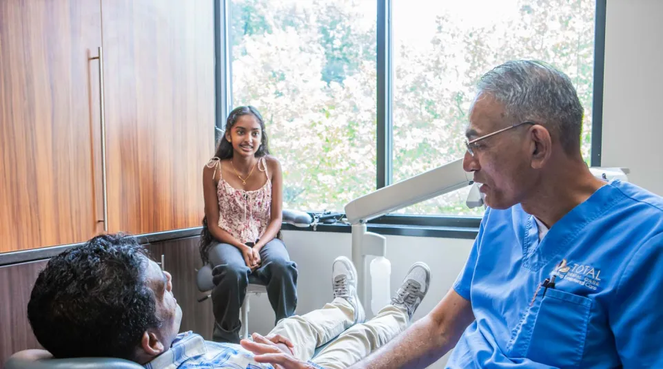 Dentist in blue scrubs talking to patient in dental chair with female companion in bright clinic room.