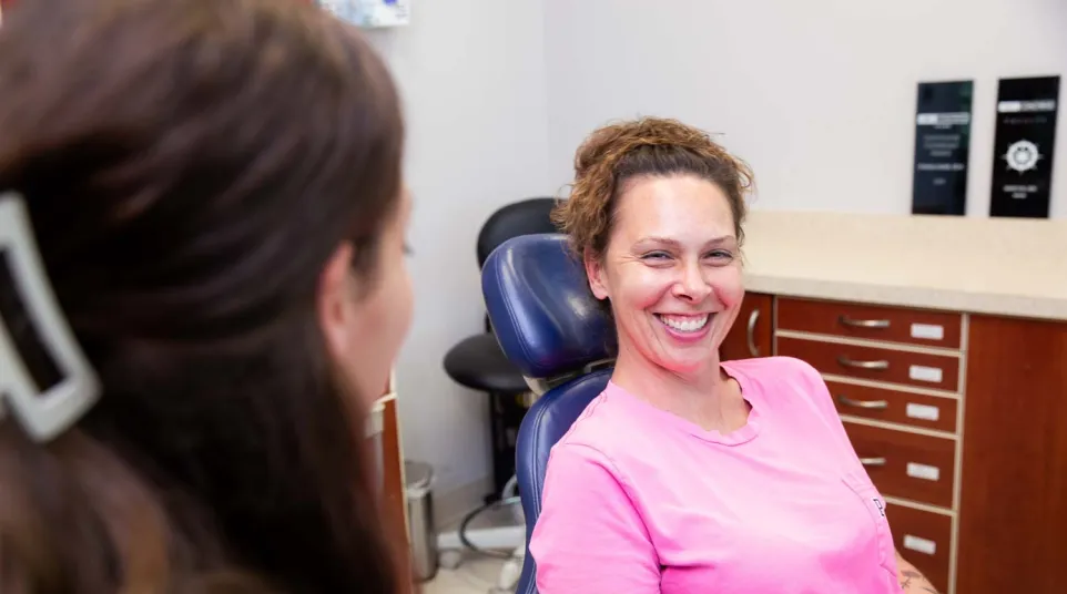 Smiling woman in a pink shirt sitting in a dental chair talking to a dentist in a clinic.