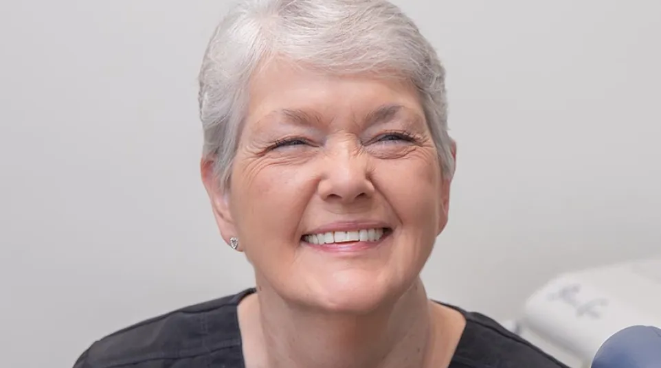Smiling elderly woman with gray hair and black top in a medical or dental office setting.