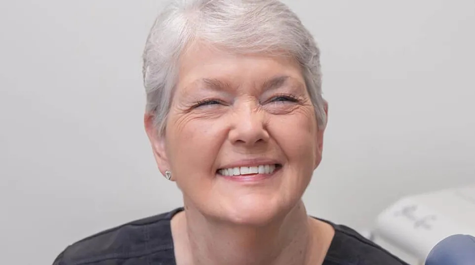 Smiling elderly woman with gray hair and black top in a medical or dental office setting.