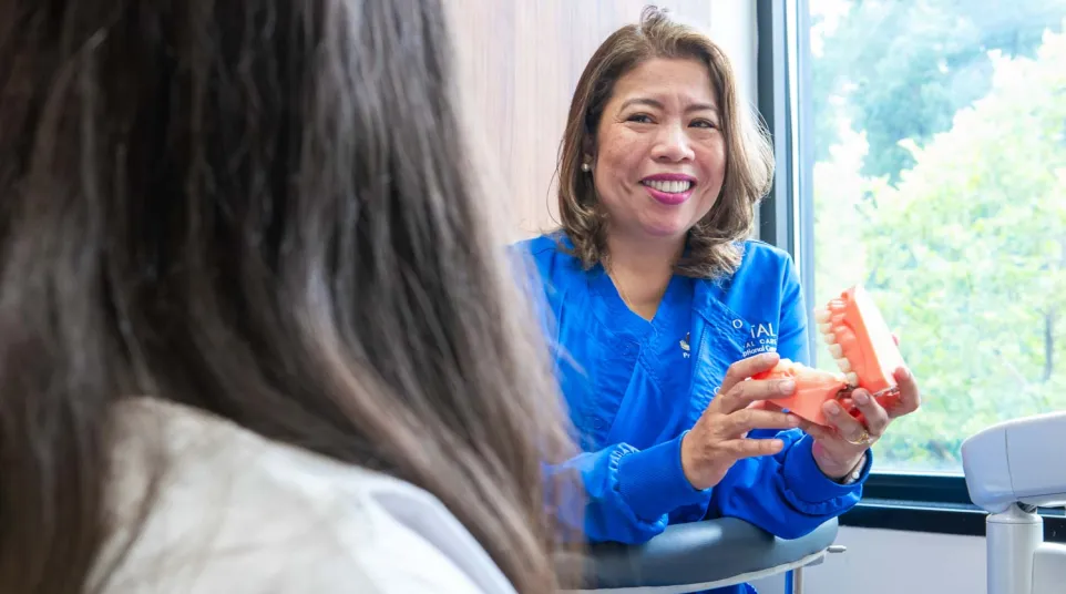 Dentist in blue scrubs smiling and showing a dental model to a patient in a consultation room.