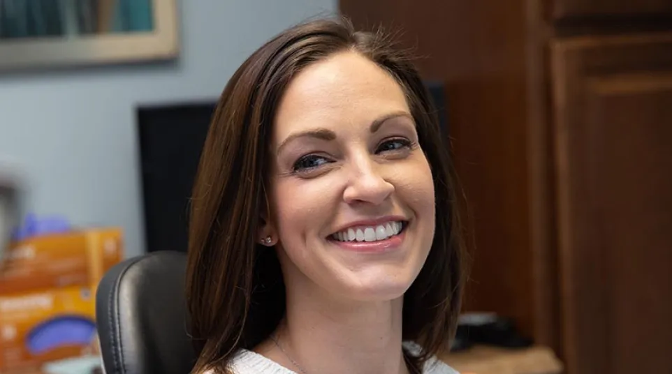 Smiling woman with long brown hair wearing a white sleeveless top sitting in an office chair indoors.